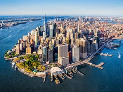 Aerial view of Lower Manhattan, New York City, showing skyscrapers, waterfront, piers, and boats on the surrounding rivers under a clear blue sky.