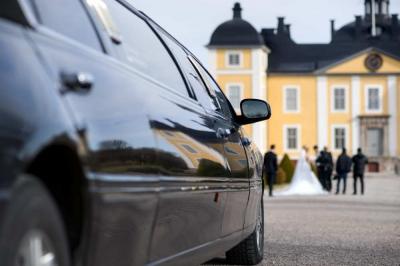 A black limousine is parked in front of a yellow mansion, with a group of people, including a bride in a white dress, gathered in the background.