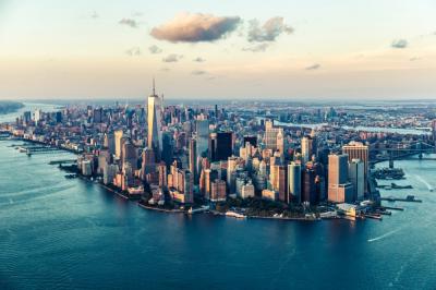Aerial view of Lower Manhattan’s skyline with tall skyscrapers, surrounding rivers, and a partly cloudy sky at sunset.