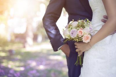 A bride and groom stand together outdoors. The bride holds a bouquet of pastel flowers while wearing a white lace dress, and the groom wears a dark suit. Sunlight filters through the background.