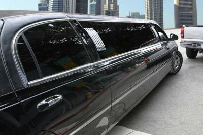 A shiny black limousine is parked on a city street with tall buildings in the background and a white pickup truck nearby.
