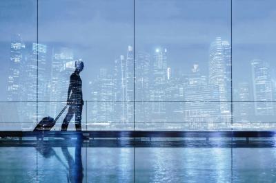 A person with a suitcase walks through an airport terminal, silhouetted against large windows showing a city skyline at dusk. Reflections are visible on the shiny floor.