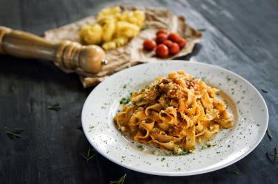 A white plate of tagliatelle pasta with sauce and grated cheese sits on a dark table. In the background, gnocchi, cherry tomatoes, and a wooden pepper grinder are on parchment paper.