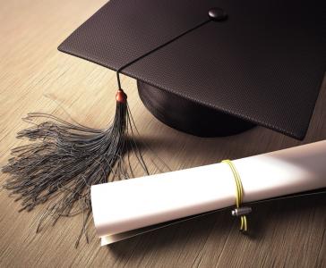 A black graduation cap with a tassel is placed next to a rolled-up diploma tied with a yellow string on a light wooden surface.