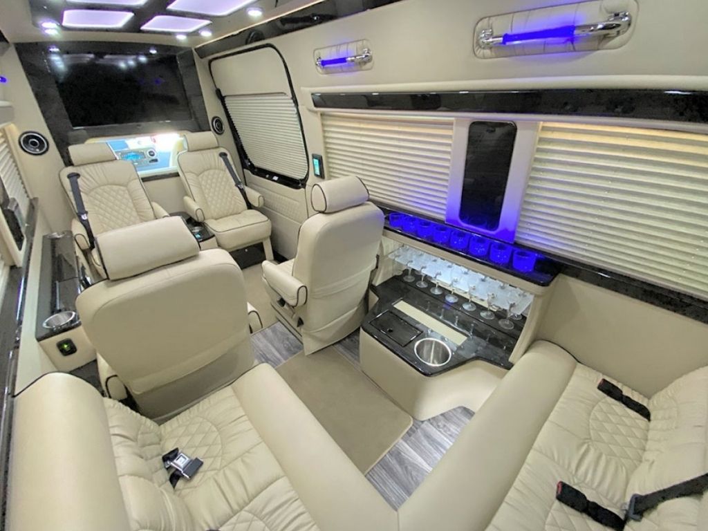 Luxurious van interior with cream leather seats, LED lighting, cup holders, and window shades. The spacious layout includes lounge seating and a control panel, creating a comfortable, modern travel environment.