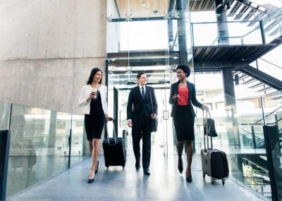 Three business professionals, two women and one man, walk through a modern building with luggage, dressed in formal attire, appearing to converse as they head to their destination.