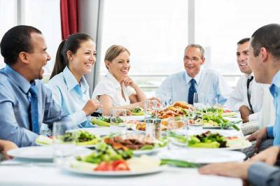 Six people in business attire sit around a table, smiling and talking, with plates of food and salads in front of them in a bright, modern setting.