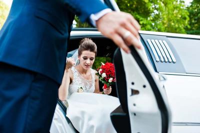 A bride holding a bouquet of red flowers steps out of a white car, assisted by a person in a suit, with green trees visible in the background.