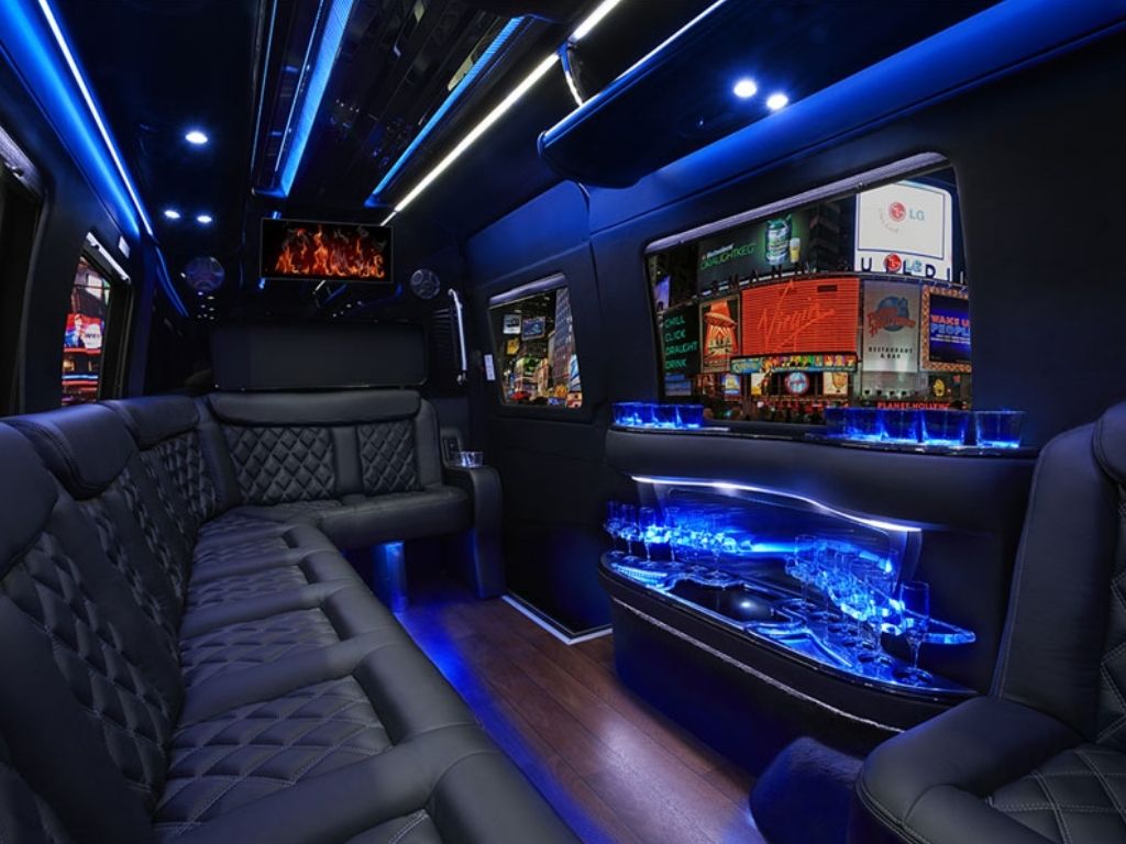 Luxurious limo interior with black leather seats, blue LED lighting, hardwood floors, a mirrored bar with glasses, and TV screens displaying colorful images and a fireplace scene.