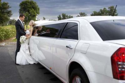 A bride and groom in wedding attire stand beside a white stretch limousine on a cloudy day, gazing at each other and smiling.
