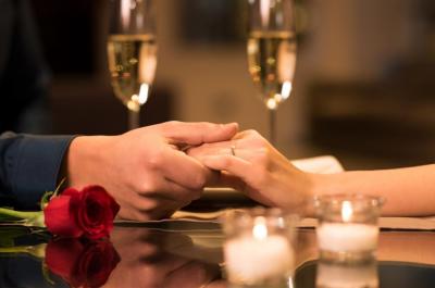 Two people holding hands across a table with a red rose, two glasses of champagne, and small candles, suggesting a romantic dinner setting.